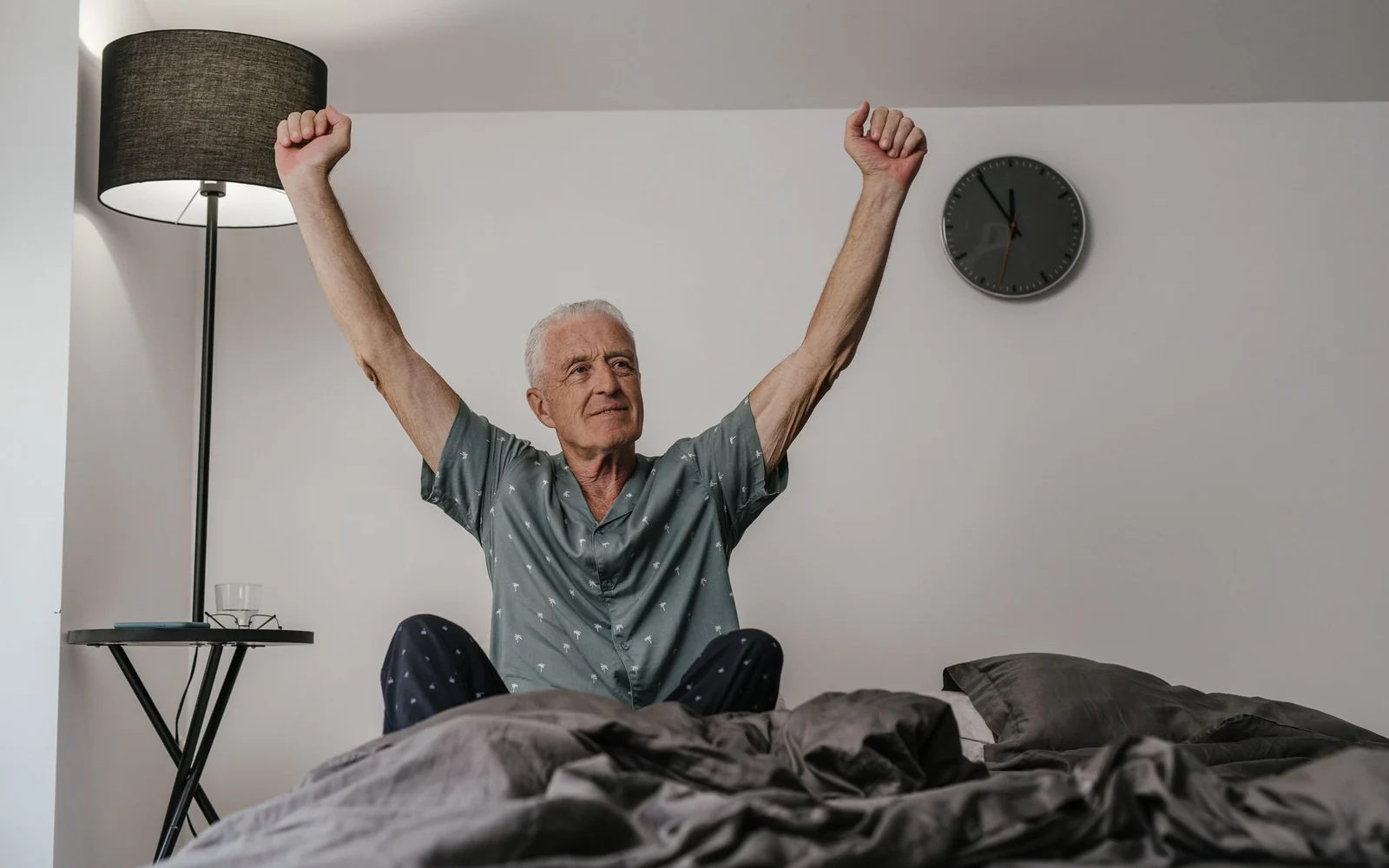 An older man in long patterned pyjamas stretches his arms while sitting in bed.