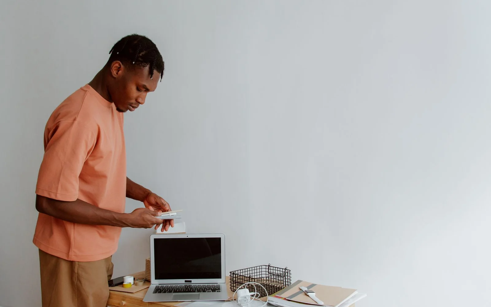 A man in an orange T-shirt and brown trousers stands beside a desk and open laptop.