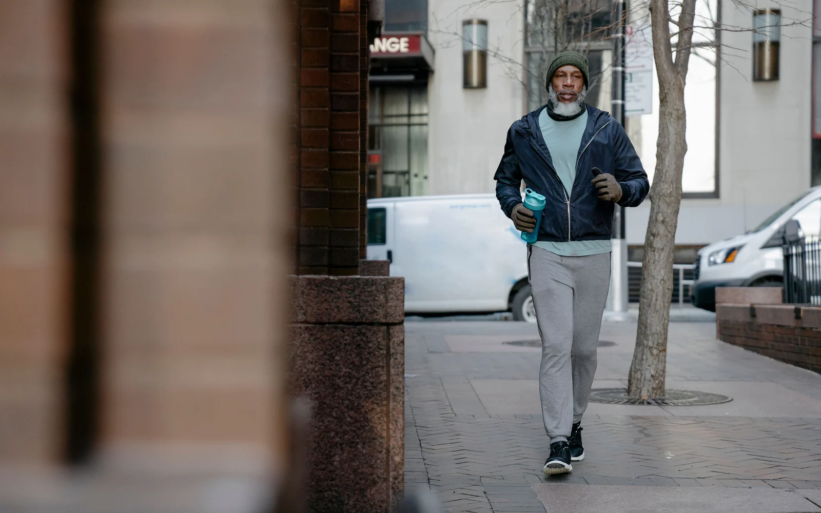 An older man in full-length grey trousers and a navy jacket moves along a city pavement holding a water bottle.